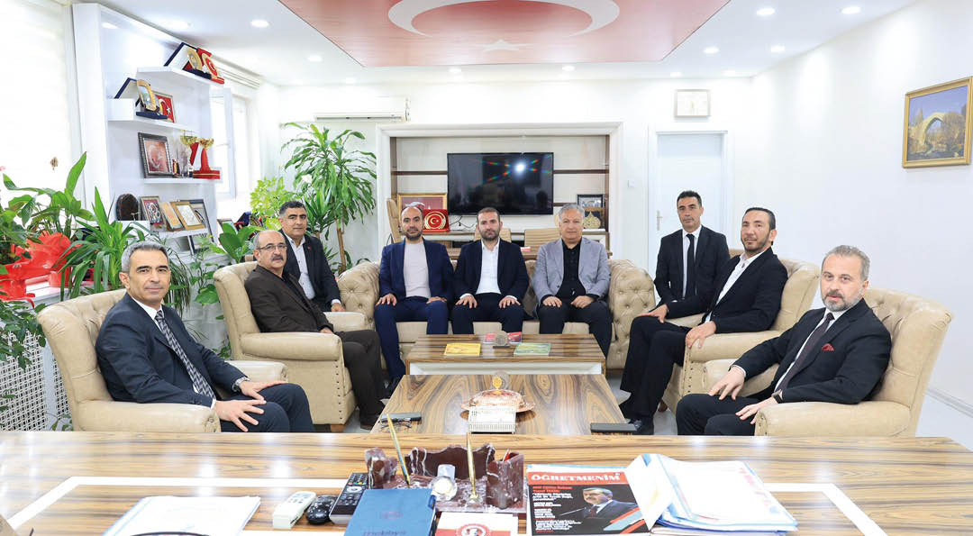 Seven people dressed in formal attire are seated on beige sofas around a wooden coffee table in a well-lit office with plants, shelves, and a television on the wall. Description generated by AI