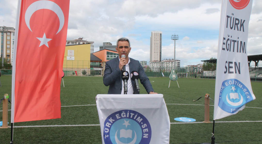 A person in a suit is speaking into a microphone behind a podium with a banner that reads “T RK E T M SEN 1992" on a sports field with two flags displaying the same text on either side.  Description generated by AI