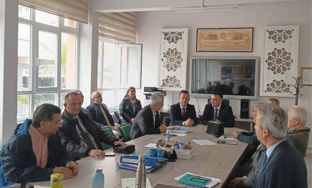 A group of adults in formal and casual clothing are seated around a conference table in a well-lit office with large windows, engaged in a meeting. Description generated by AI
