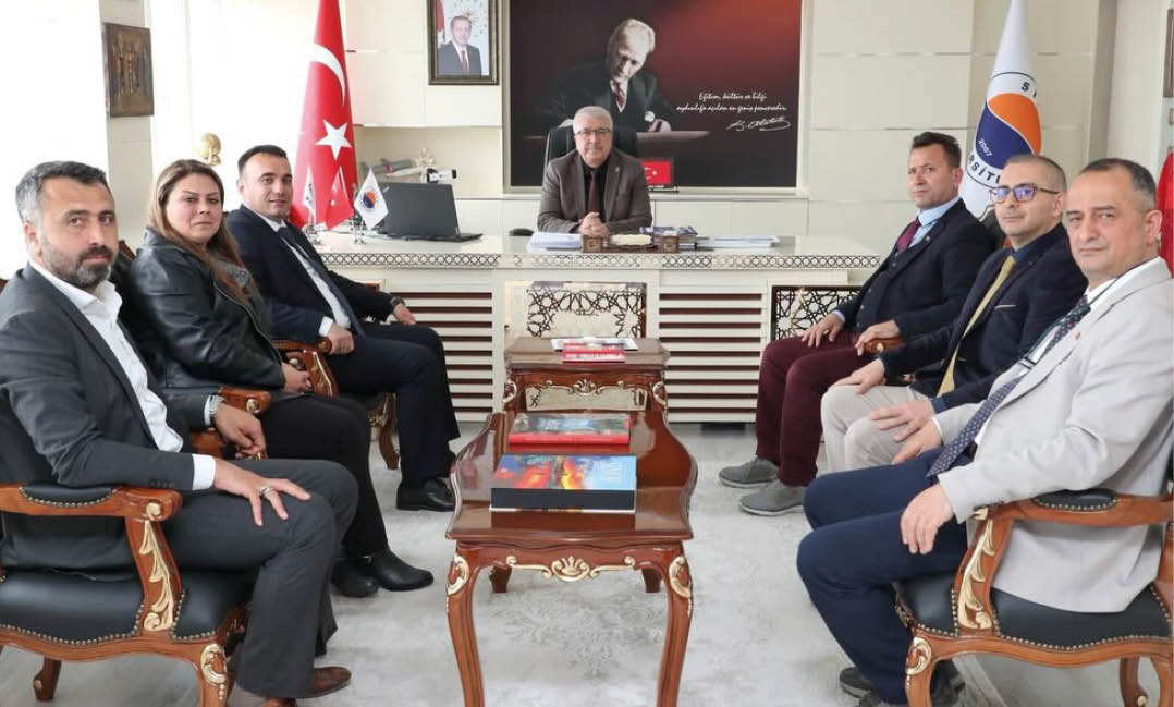Seven people in formal attire are seated in a meeting room with a person at a desk in the center, Turkish flags and a portrait with a quote are displayed on the wall behind. Description generated by AI