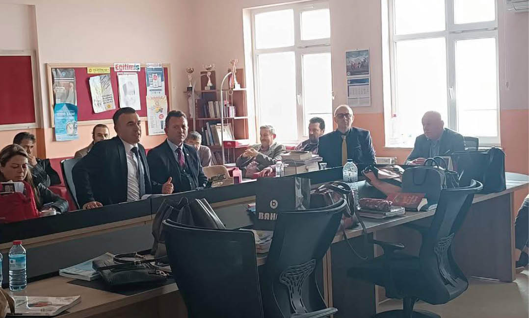 People in formal attire are seated and standing around desks in a well-lit office or meeting room with large windows and various office supplies. Description generated by AI