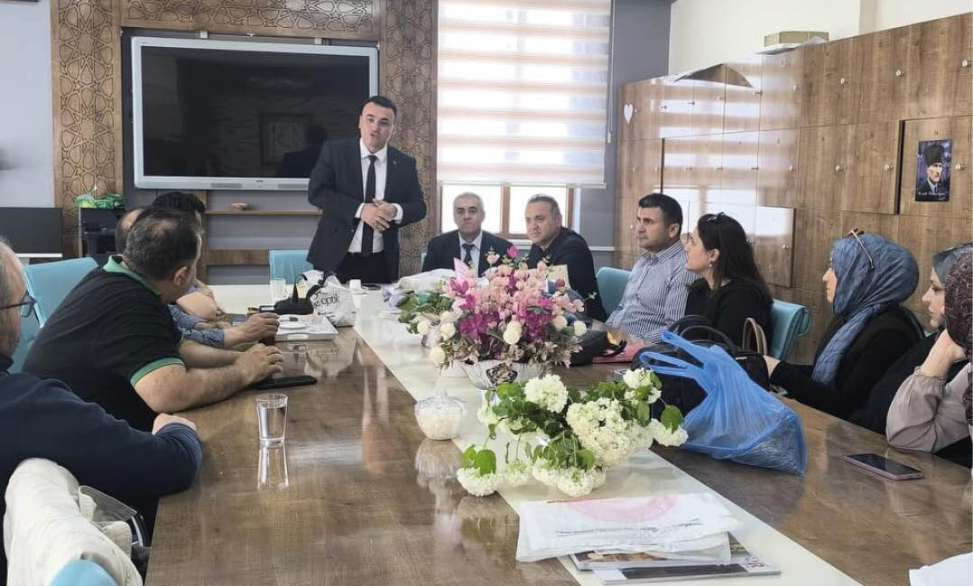 People dressed in formal and casual clothing are seated around a conference table with floral centerpieces while one person stands speaking in a modern office setting. Description generated by AI