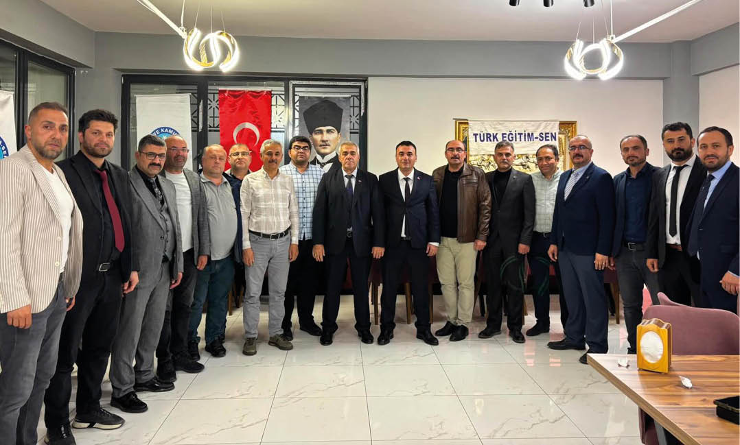 A group of adults dressed in formal and casual clothing stand together indoors in front of flags and a banner with Turkish text. Description generated by AI