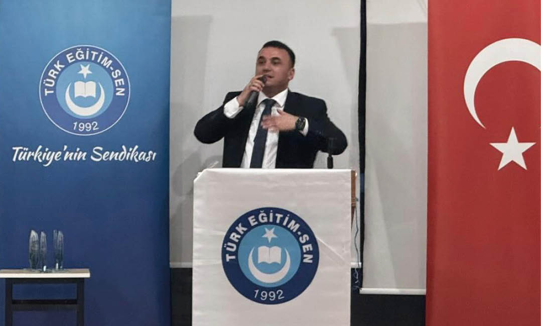A person in a suit is speaking at a podium with a microphone between two banners, one blue with the text “T RK E T M-SEN 1992 T rkiye'nin Sendikas " and the other red with a white star and crescent.  Description generated by AI
