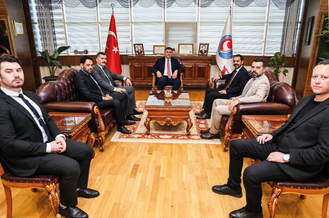 Six people in formal attire are seated in a well-decorated office with flags, plants, and framed certificates, engaged in a meeting around a central coffee table. Description generated by AI
