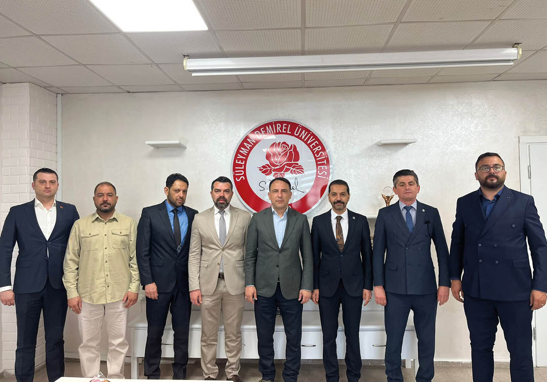 Five people dressed in formal suits stand in front of a wall with a large circular emblem featuring a rose and the text “S leyman Demirel niversitesi."  Description generated by AI