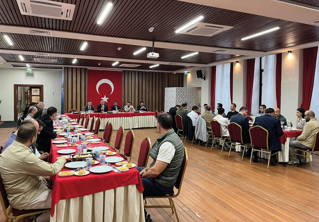 Adults are seated around tables in a meeting room with a large flag featuring a crescent and star hanging on the wall. Description generated by AI