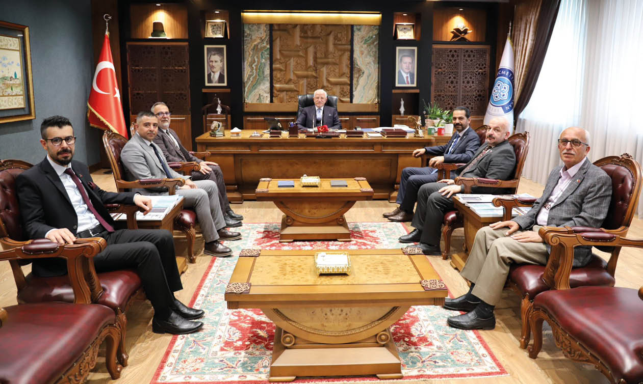 Several people in formal attire are seated around a large wooden desk in an ornate office setting with decorative rugs and plants. Description generated by AI
