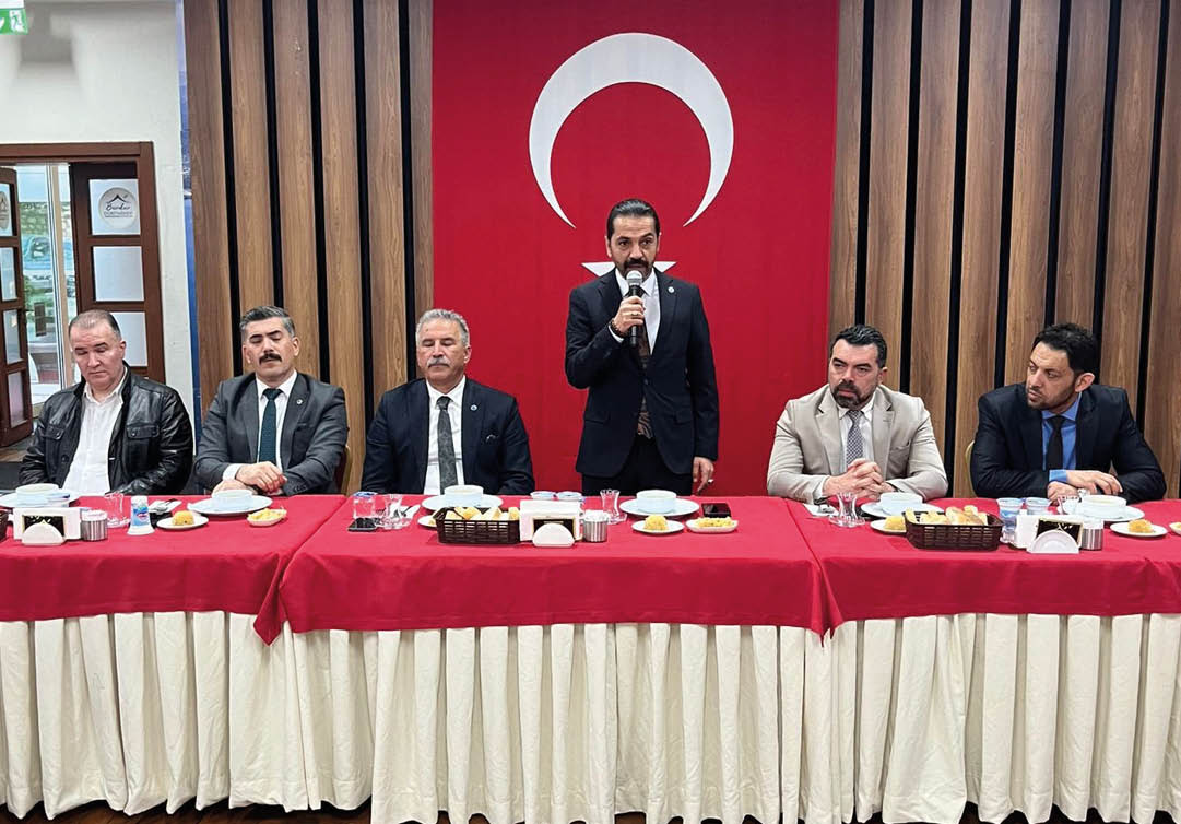 Four adults in formal attire sit at a table with red tablecloths and white skirts while one adult in a dark suit stands speaking into a microphone in front of a large red flag with a white crescent and star. Description generated by AI