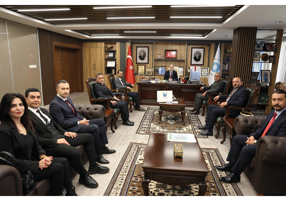 Several people in formal attire are seated in a wood-paneled office with flags, portraits, and a large desk where one person is sitting behind it. Description generated by AI