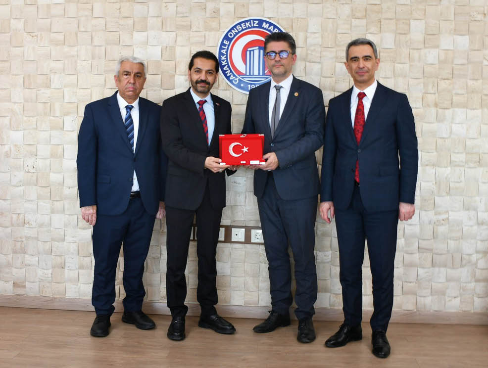 Four people in suits stand indoors with two in the center holding a red box featuring a white crescent and star symbol. Description generated by AI