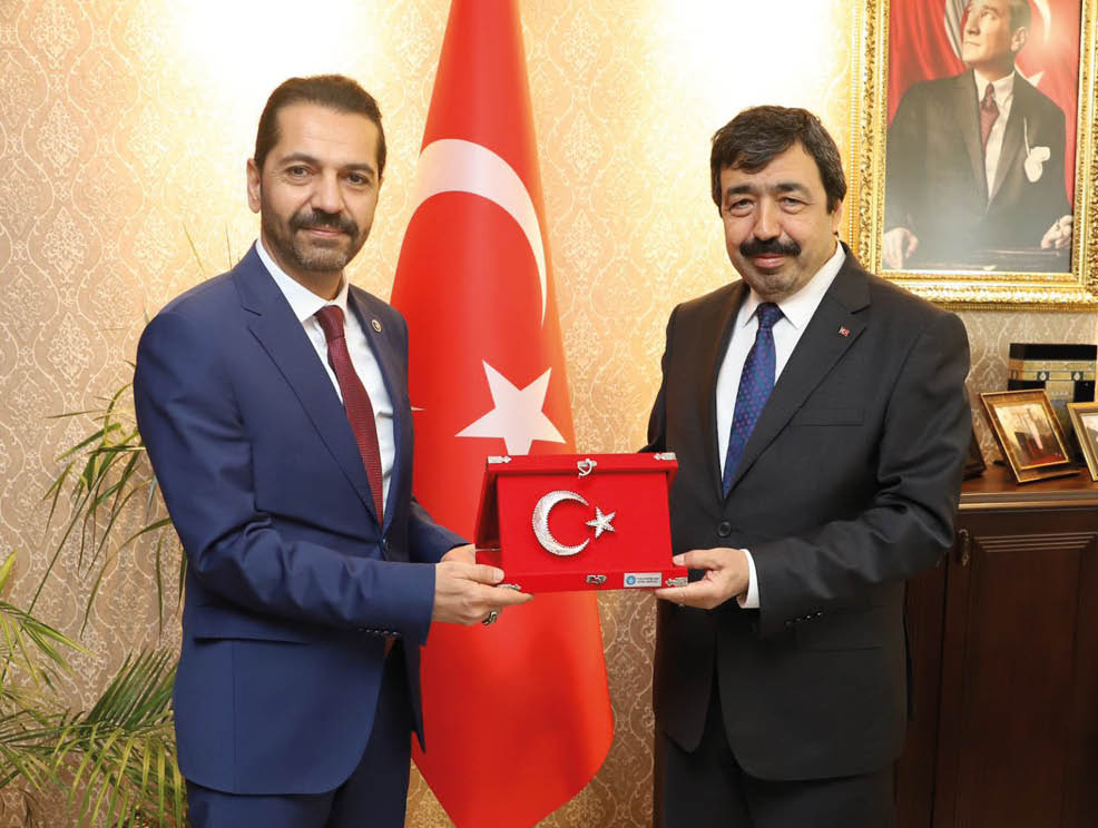 Two adults in suits stand indoors holding a red plaque featuring a crescent and star symbol with a Turkish flag in the background. Description generated by AI