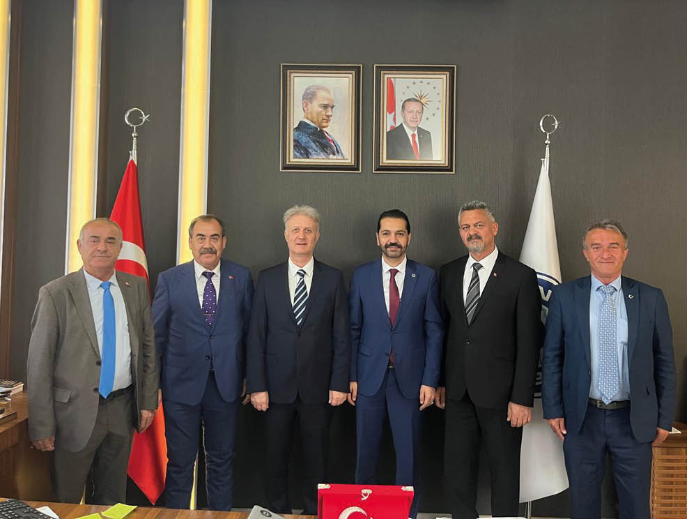 Six adults in formal suits stand side by side in front of a dark wall with two framed portraits and flags on either side. Description generated by AI