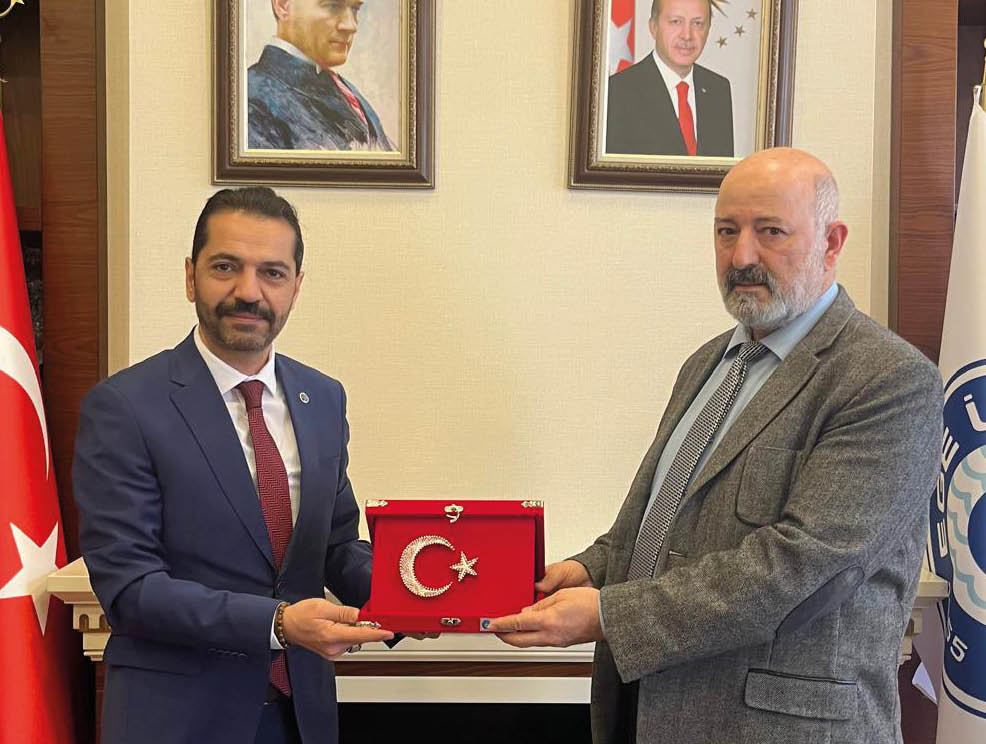 Two people in formal attire are standing indoors holding a red plaque featuring a crescent and star symbol between them. Description generated by AI