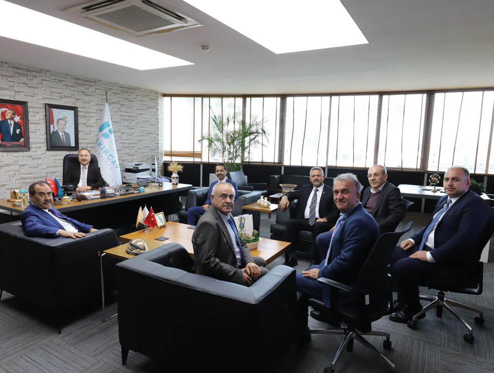 A group of people in business attire are seated and engaged in a meeting in a modern office with large windows and a flag in the background. Description generated by AI