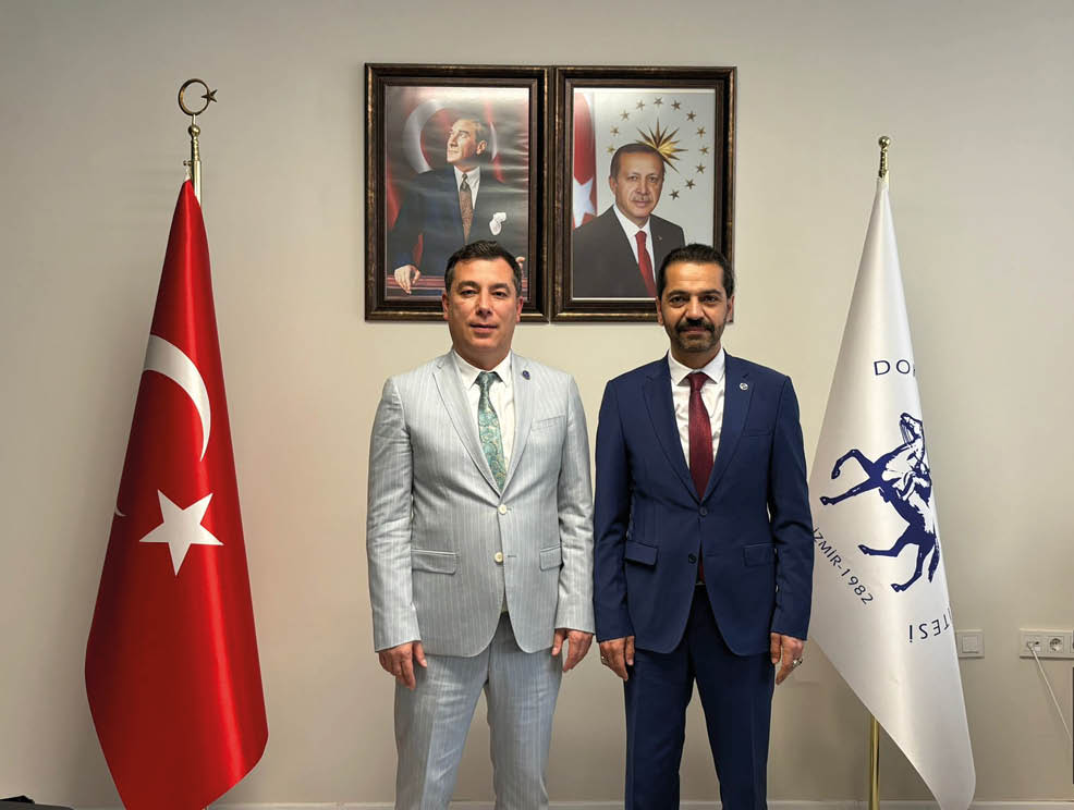 Two adults wearing formal suits stand side by side in front of a wall with framed portraits. Description generated by AI