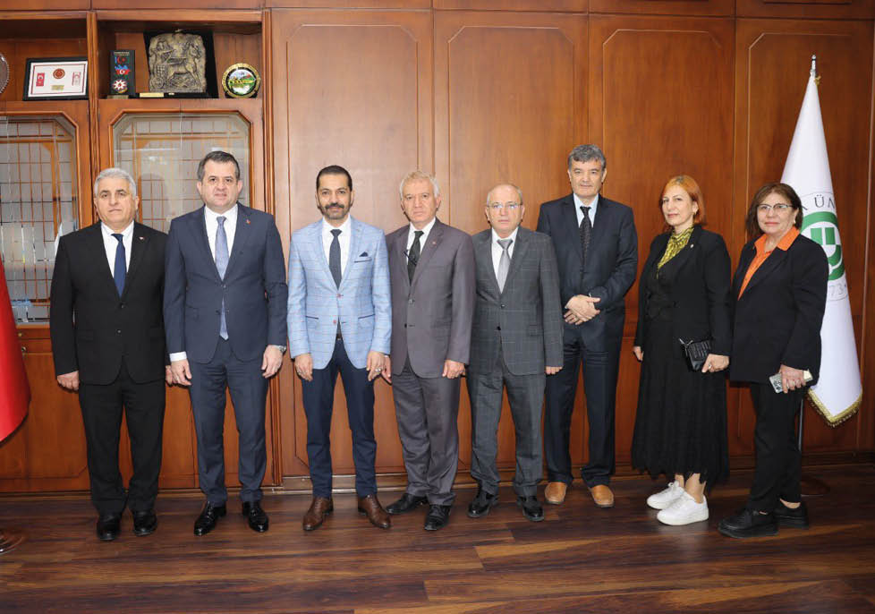 Seven adults dressed in formal and semi-formal attire stand side by side in front of a wooden wall with a display cabinet on the left. Description generated by AI