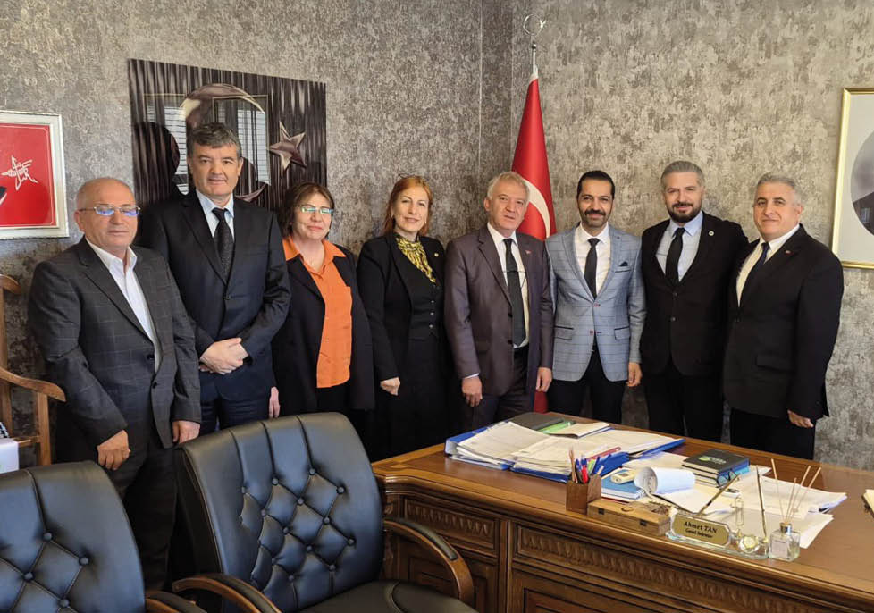 Seven people dressed in formal attire stand behind a wooden desk covered with papers, folders, and office supplies in an office setting with a flag and framed portrait on the wall. Description generated by AI