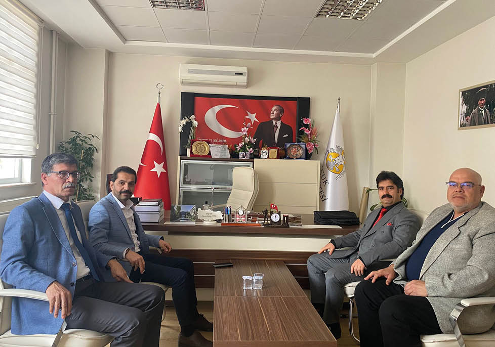An office desk with two people seated on either side, a large Turkish flag and a portrait behind the desk, along with various plaques, flowers, and office supplies on the desk and shelves. Description generated by AI