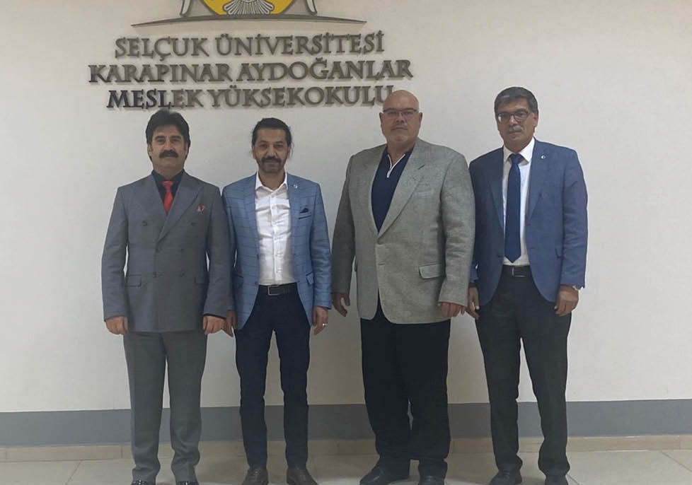 Four adults dressed in formal attire stand side by side in front of a wall with the text “Sel uk niversitesi Karap nar Aydo anlar Meslek Y ksekokulu."  Description generated by AI