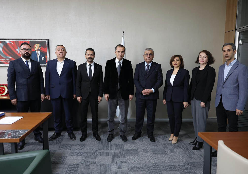 Seven adults dressed in formal business attire stand side by side in an office setting with a framed portrait and a flag in the background. Description generated by AI