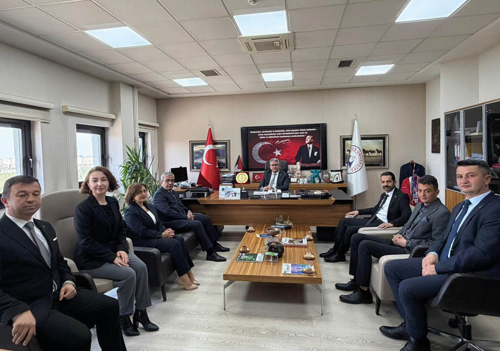 A group of people in formal attire are seated in an office around a large desk with flags, plaques, and a framed portrait of a historical figure with a flag in the background. Description generated by AI