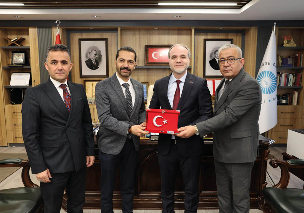Four people in suits stand indoors holding a red plaque with a white crescent and star symbol, with framed pictures and a flag in the background. Description generated by AI