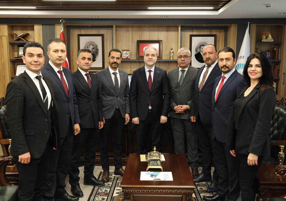Seven adults dressed in formal suits stand closely together in an office setting with framed portraits and decorative items on wooden shelves behind them. Description generated by AI