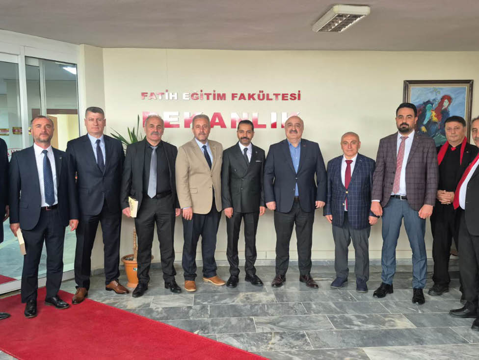 A group of nine adults dressed in formal business attire stand side by side indoors in front of a wall with red text and a potted plant. Description generated by AI