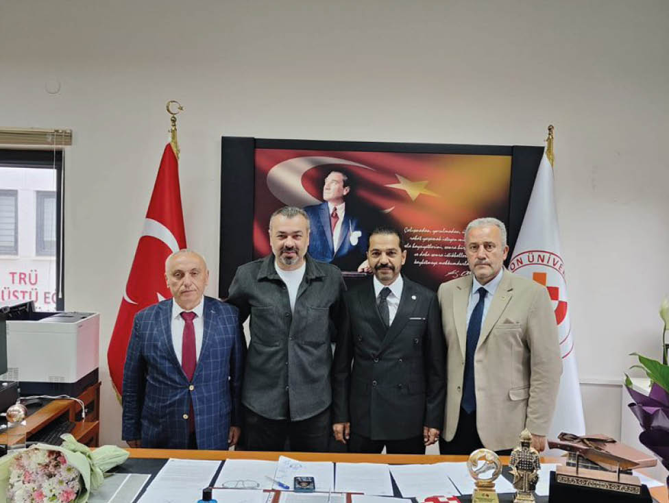 Four adults dressed in formal and semi-formal attire stand behind a desk with papers and awards in an office decorated with two flags and a large portrait featuring a person and a flag. Description generated by AI