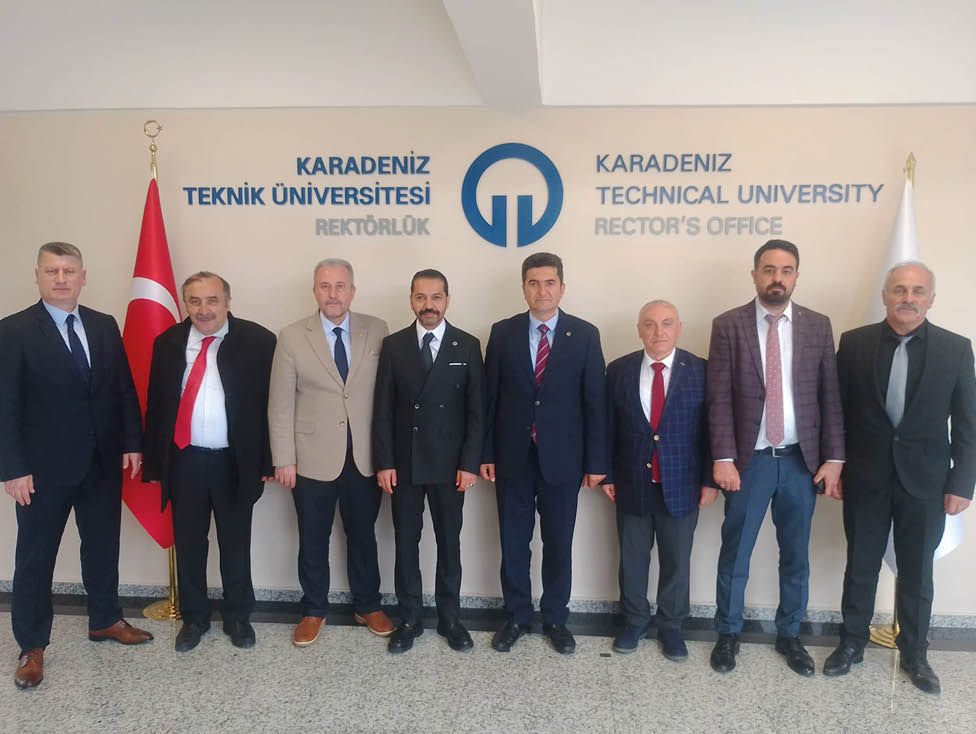 Five adults dressed in formal attire stand side by side in front of a wall with text and a logo related to a university rector's office. Description generated by AI