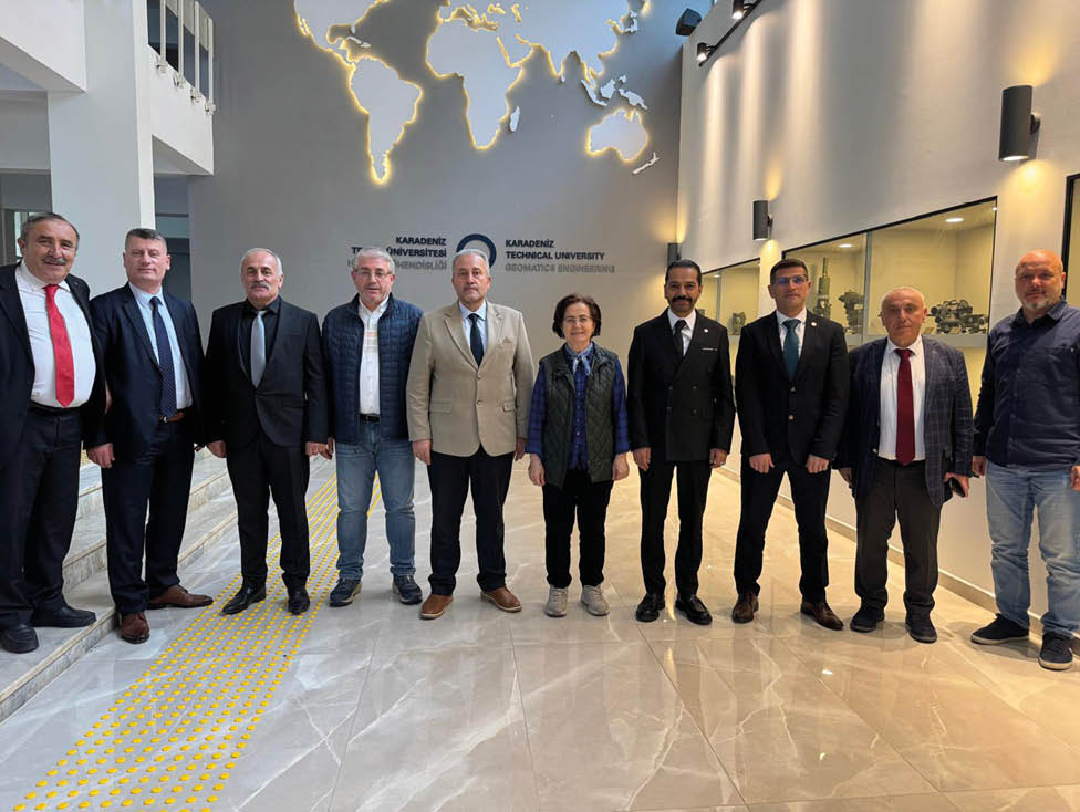 Six adults dressed in business and casual attire stand side by side in a modern indoor setting with a wall displaying the name Karadeniz Technical University behind them. Description generated by AI