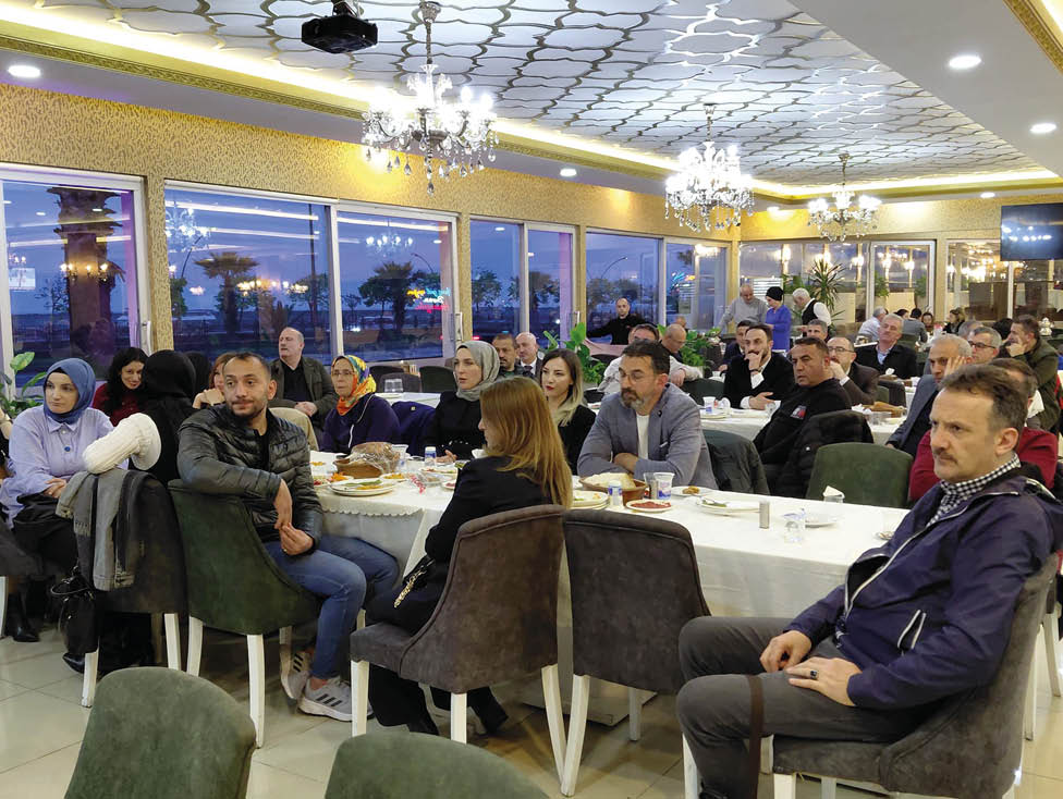 People are seated around tables with food and drinks in a well-lit indoor dining area with large windows showing an evening outdoor scene. Description generated by AI