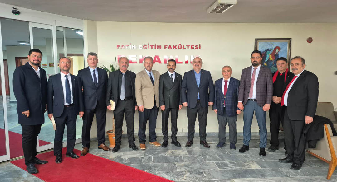 A group of nine adults dressed in formal business attire stand side by side indoors in front of a wall with red text and a potted plant. Description generated by AI