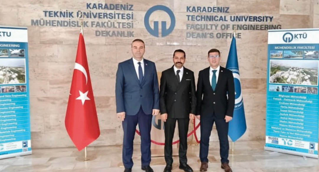 Three adults in formal suits stand side by side in front of a wall with the Karadeniz Technical University Faculty of Engineering Dean's Office signage and two flags. Description generated by AI