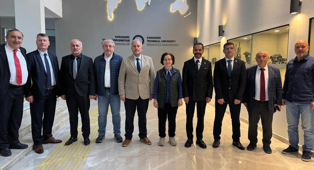 Six adults dressed in business and casual attire stand side by side in a modern indoor setting with a wall displaying the name Karadeniz Technical University behind them. Description generated by AI