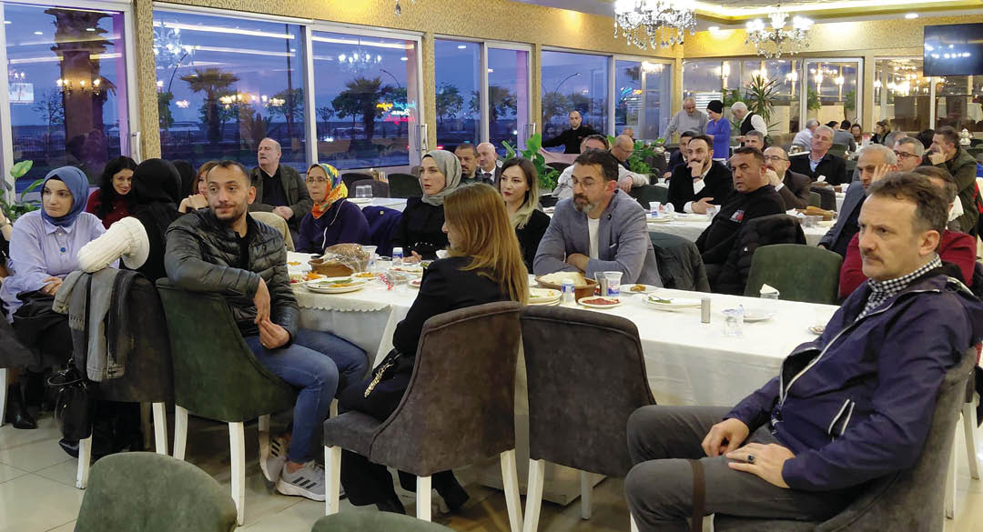 People are seated around tables with food and drinks in a well-lit indoor dining area with large windows showing an evening outdoor scene. Description generated by AI