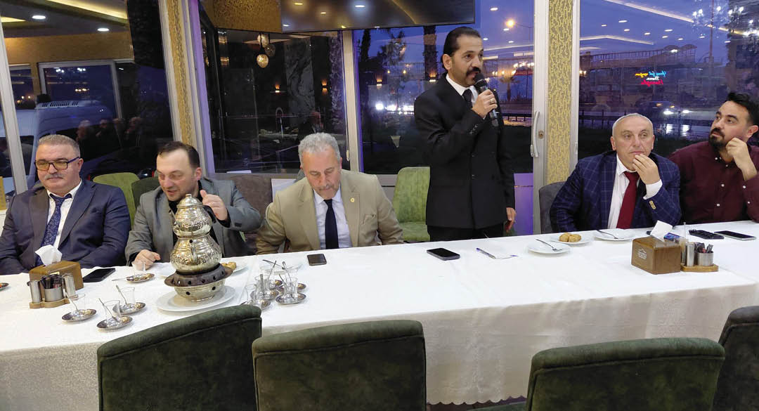 A person in a suit is standing and speaking into a microphone while another person in a beige suit is seated at a table with a white tablecloth, with phones and small dishes on the table. Description generated by AI