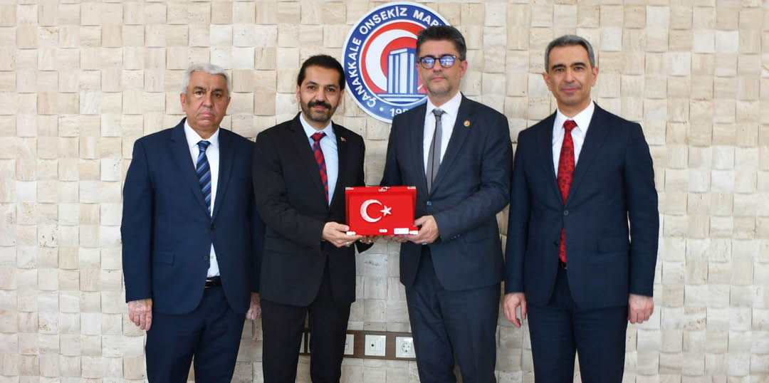 Four people in suits stand indoors with two in the center holding a red box featuring a white crescent and star symbol. Description generated by AI