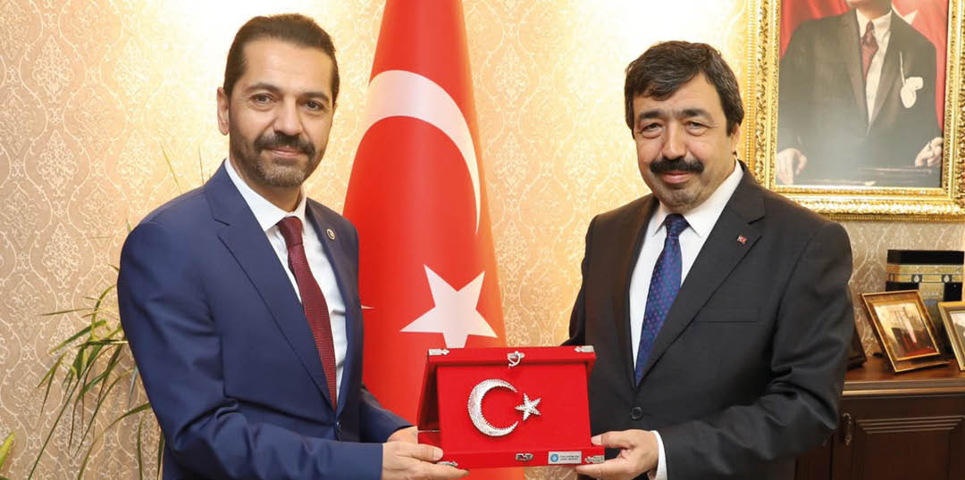 Two adults in suits stand indoors holding a red plaque featuring a crescent and star symbol with a Turkish flag in the background. Description generated by AI