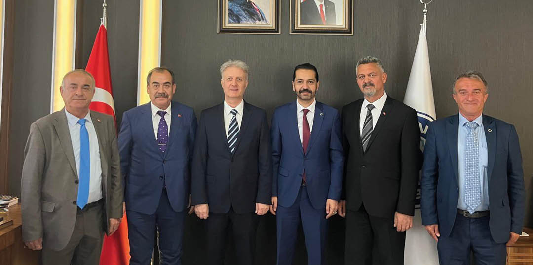 Six adults in formal suits stand side by side in front of a dark wall with two framed portraits and flags on either side. Description generated by AI