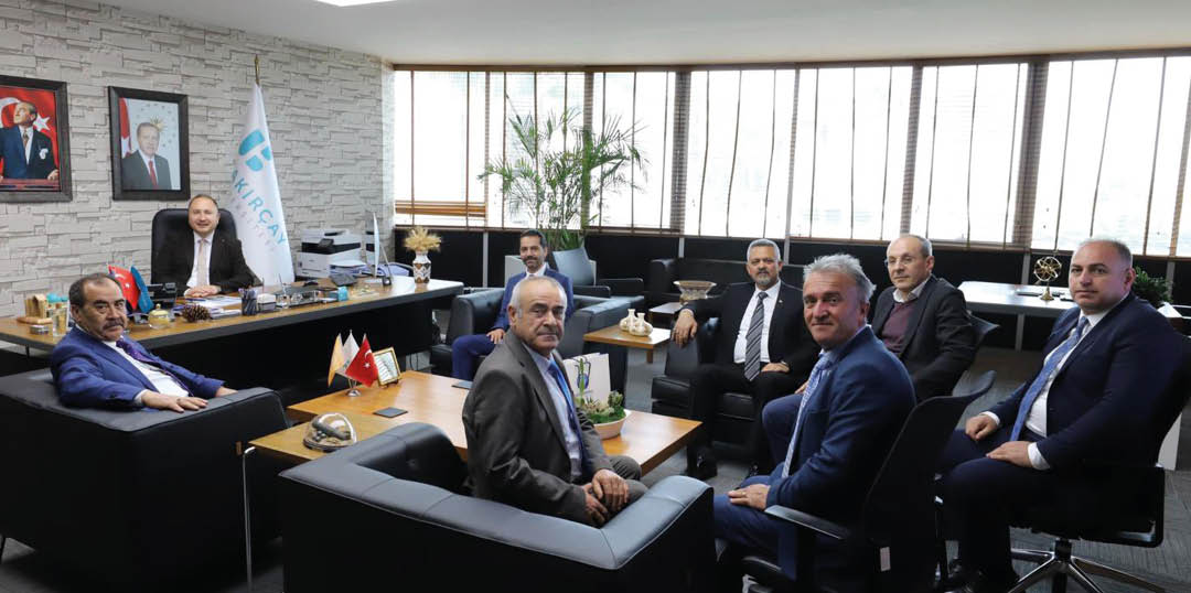 A group of people in business attire are seated and engaged in a meeting in a modern office with large windows and a flag in the background. Description generated by AI