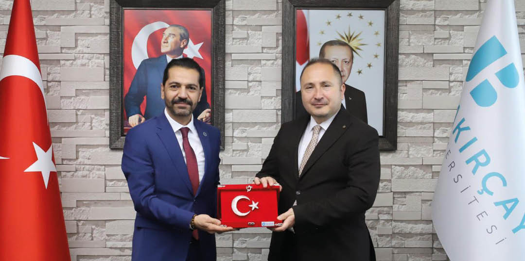 Two people in formal suits are standing indoors holding a red plaque with a white crescent and star symbol, with framed portraits and a brick-patterned wall in the background. Description generated by AI