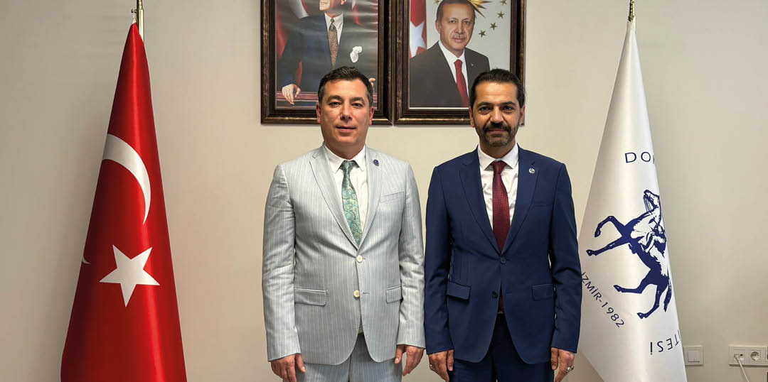 Two adults wearing formal suits stand side by side in front of a wall with framed portraits. Description generated by AI