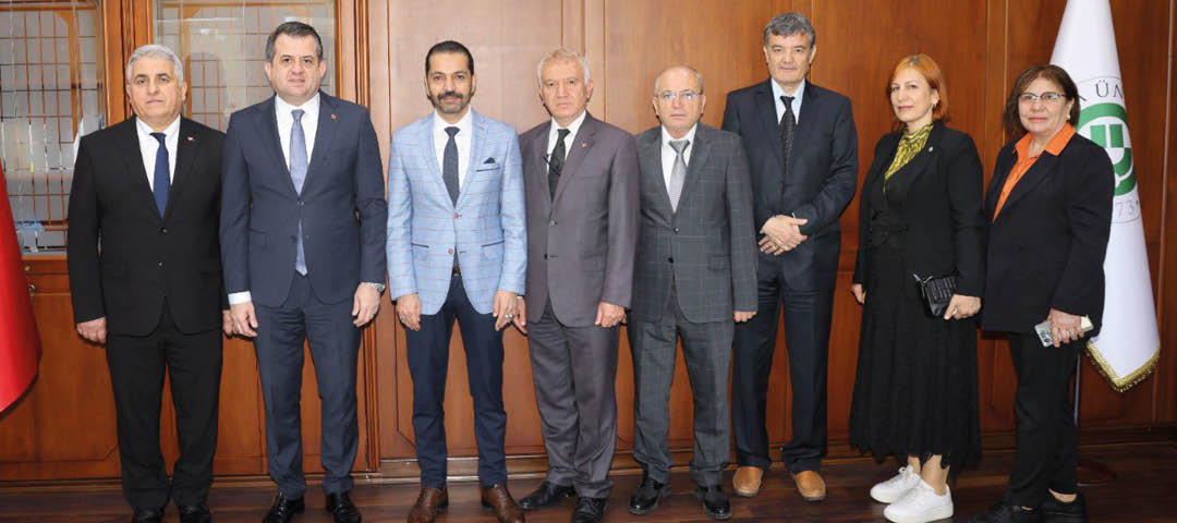 Seven adults dressed in formal and semi-formal attire stand side by side in front of a wooden wall with a display cabinet on the left. Description generated by AI