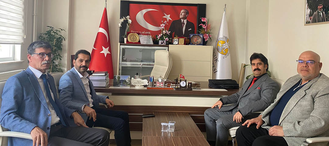 An office desk with two people seated on either side, a large Turkish flag and a portrait behind the desk, along with various plaques, flowers, and office supplies on the desk and shelves. Description generated by AI