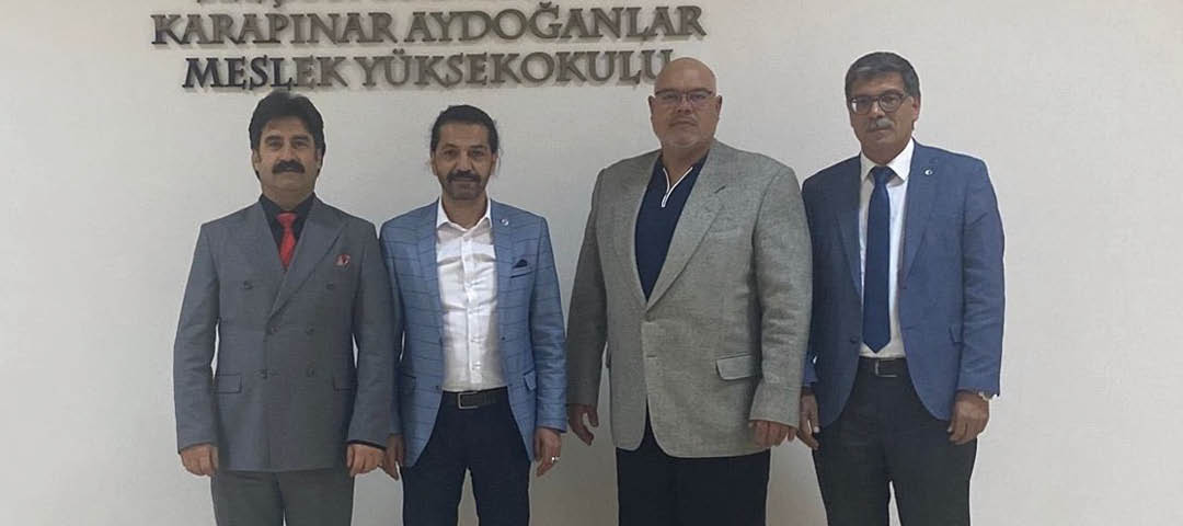 Four adults dressed in formal attire stand side by side in front of a wall with the text “Sel uk niversitesi Karap nar Aydo anlar Meslek Y ksekokulu."  Description generated by AI