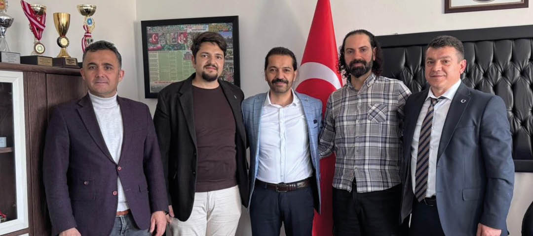 Five adults dressed in business casual and formal attire stand closely together indoors with a flag featuring a crescent and star in the background. Description generated by AI