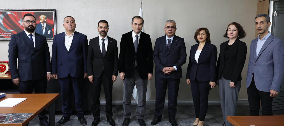 Seven adults dressed in formal business attire stand side by side in an office setting with a framed portrait and a flag in the background. Description generated by AI