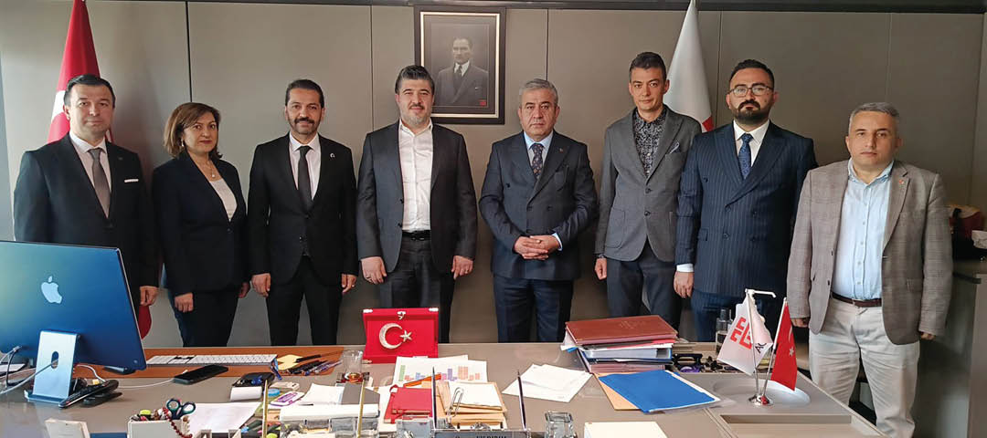 Five adults in formal attire stand behind a desk cluttered with documents, folders, a Turkish flag, and office supplies, with a framed portrait and a flag in the background. Description generated by AI